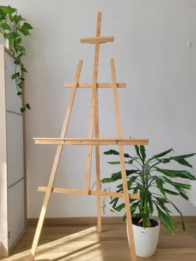 Eine leere Staffelei aus Holz steht in einem Raum neben zwei Zimmerpflanzen.