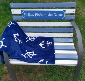 Eine blau-weiße Gartenbank mit einer maritimen Decke und einem Schild.