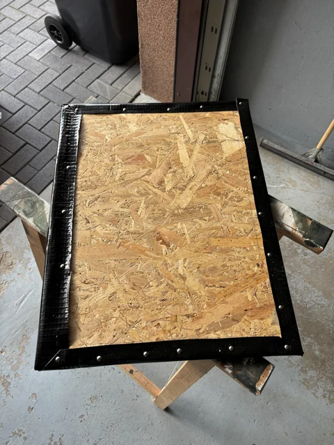 Eine OSB-Platte, eingefasst mit schwarzem Klebeband und Nieten, auf einem Holzbock.