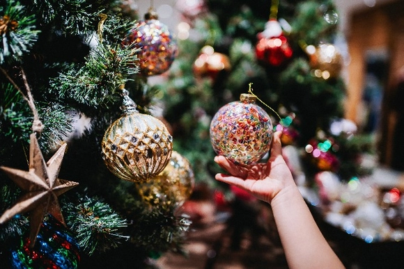 Eine Hand hält eine bunte Weihnachtskugel vor einem geschmückten Weihnachtsbaum.