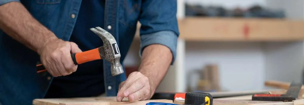 Eine Person schlägt mit einem Hammer einen Nagel in ein Holzbrett.