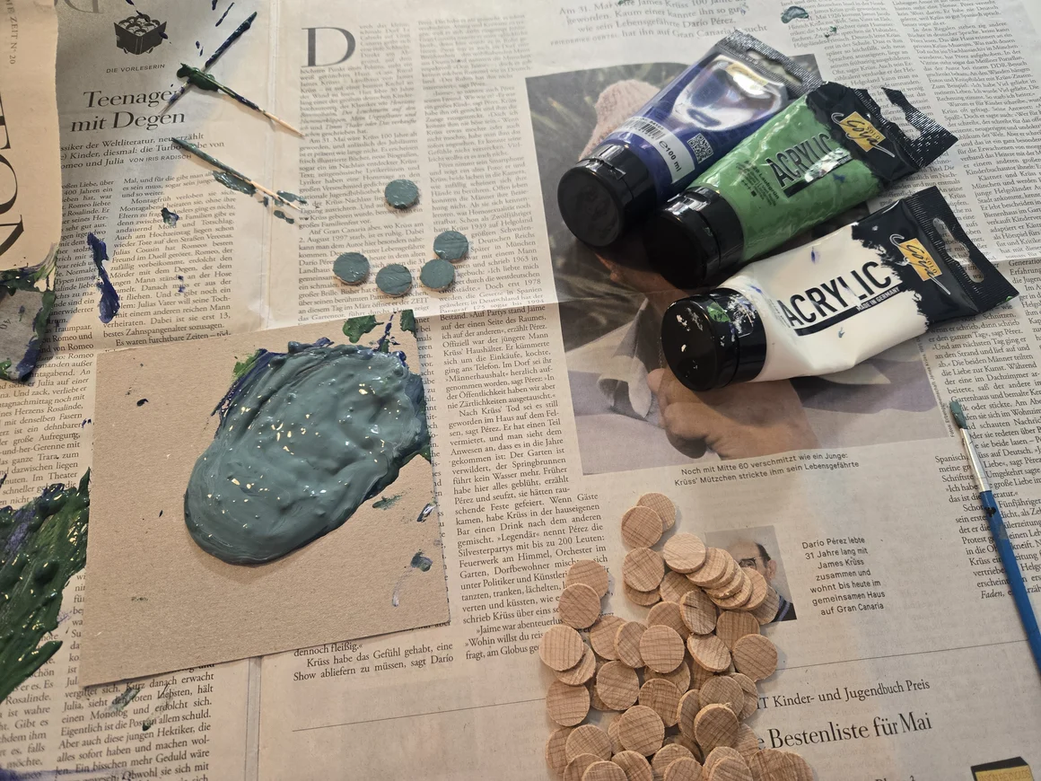 Acrylfarben, Holzplättchen und Pinsel liegen auf einer Zeitung.