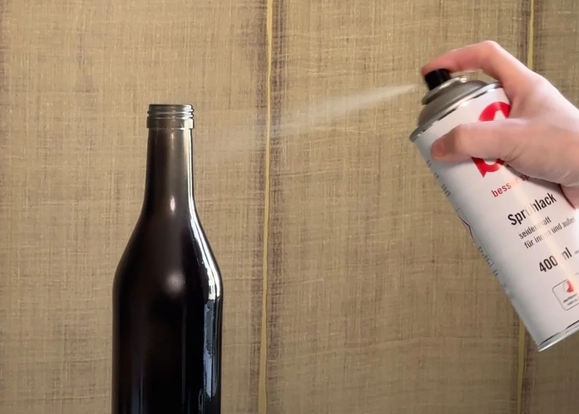 Eine Hand besprüht eine Glasflasche mit einer Sprühdose.