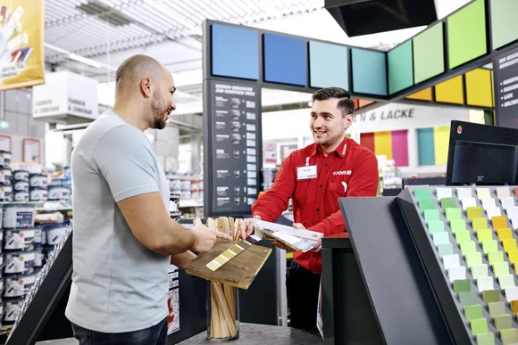 Zwei Personen im Baumarkt, eine Person reicht der anderen Farbmuster.