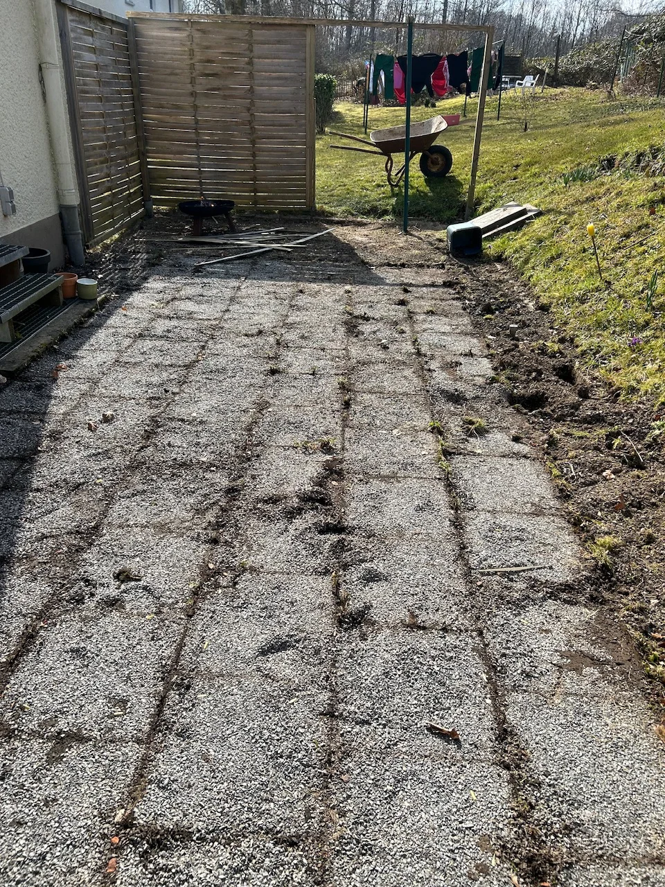 Ungepflegte Terrasse aus Waschbetonplatten mit einer Schubkarre im Hintergrund.