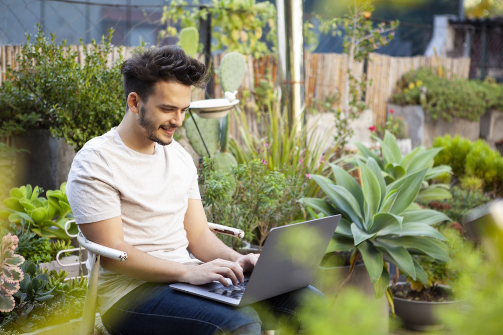 Eine lächelnde Person sitzt mit einem Laptop in einem Garten voller Pflanzen.